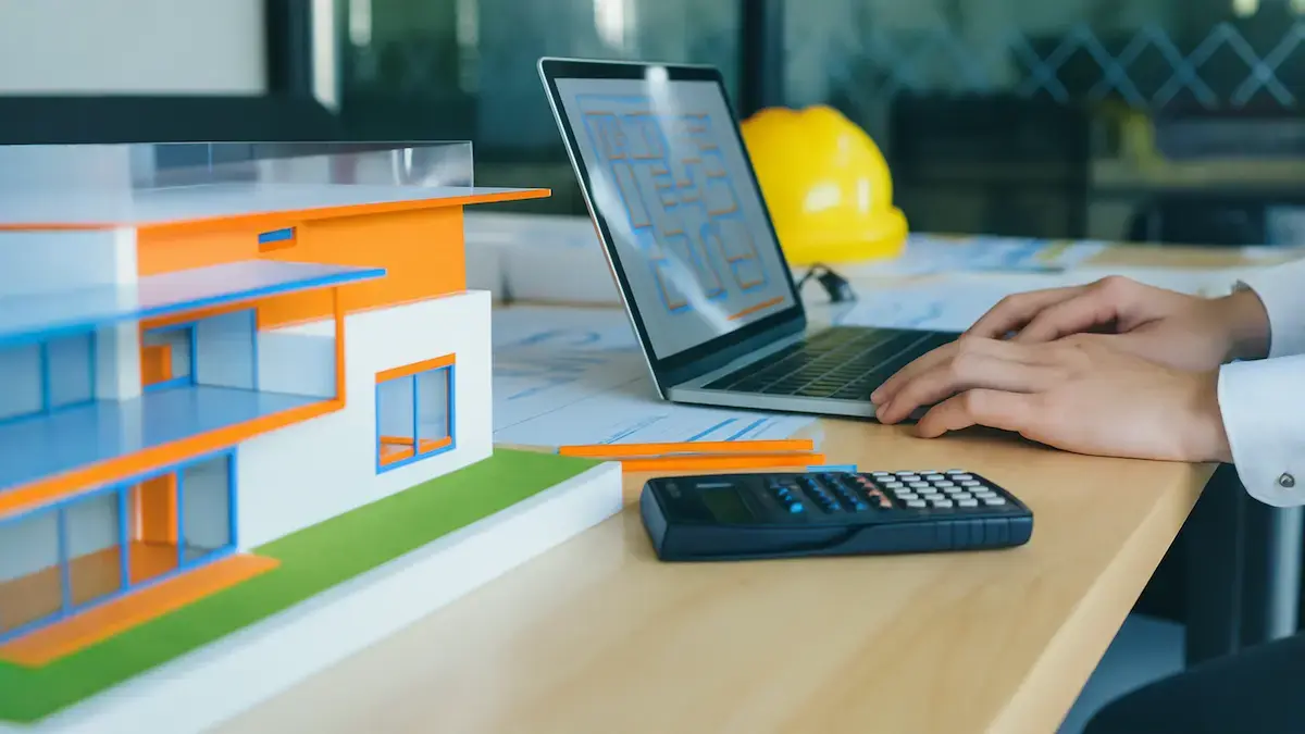 Fix and flip loan calculator setup with a scale model house, calculator, and laptop showing renovation plans on a desk.