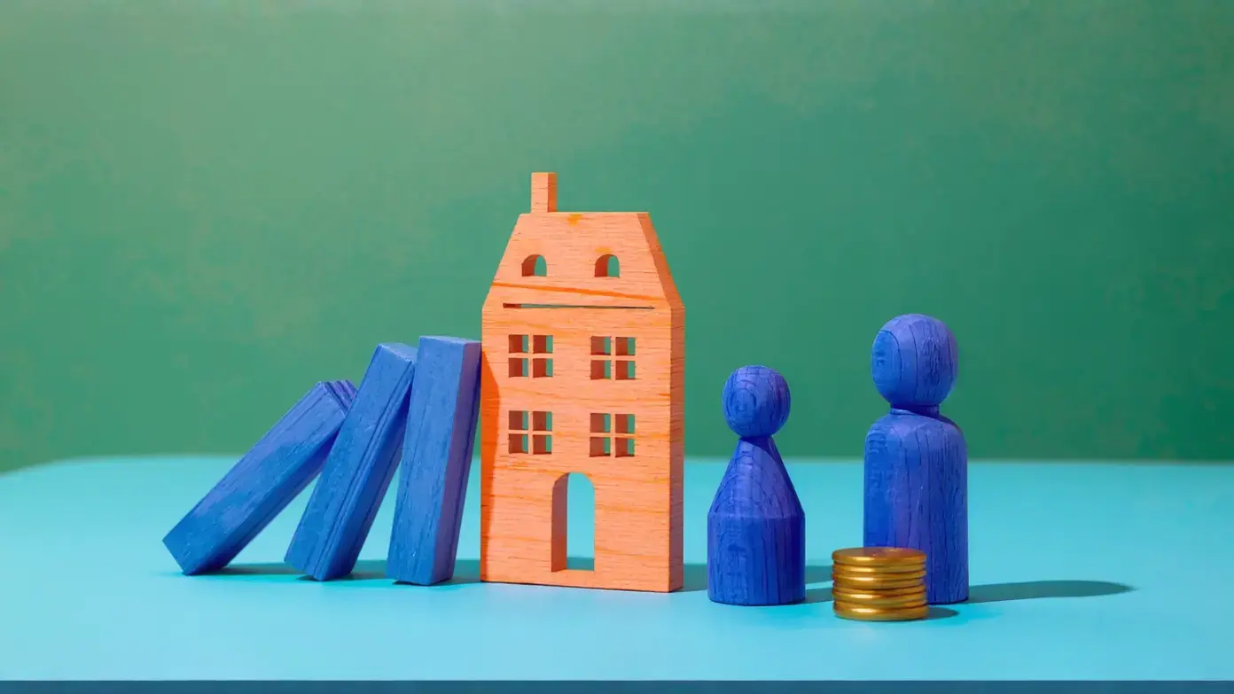 A symbolic image of private money lending risks, showing an orange wooden house with a domino chain of blue blocks leaning against it, as two wooden figures and a stack of gold coins stand by.